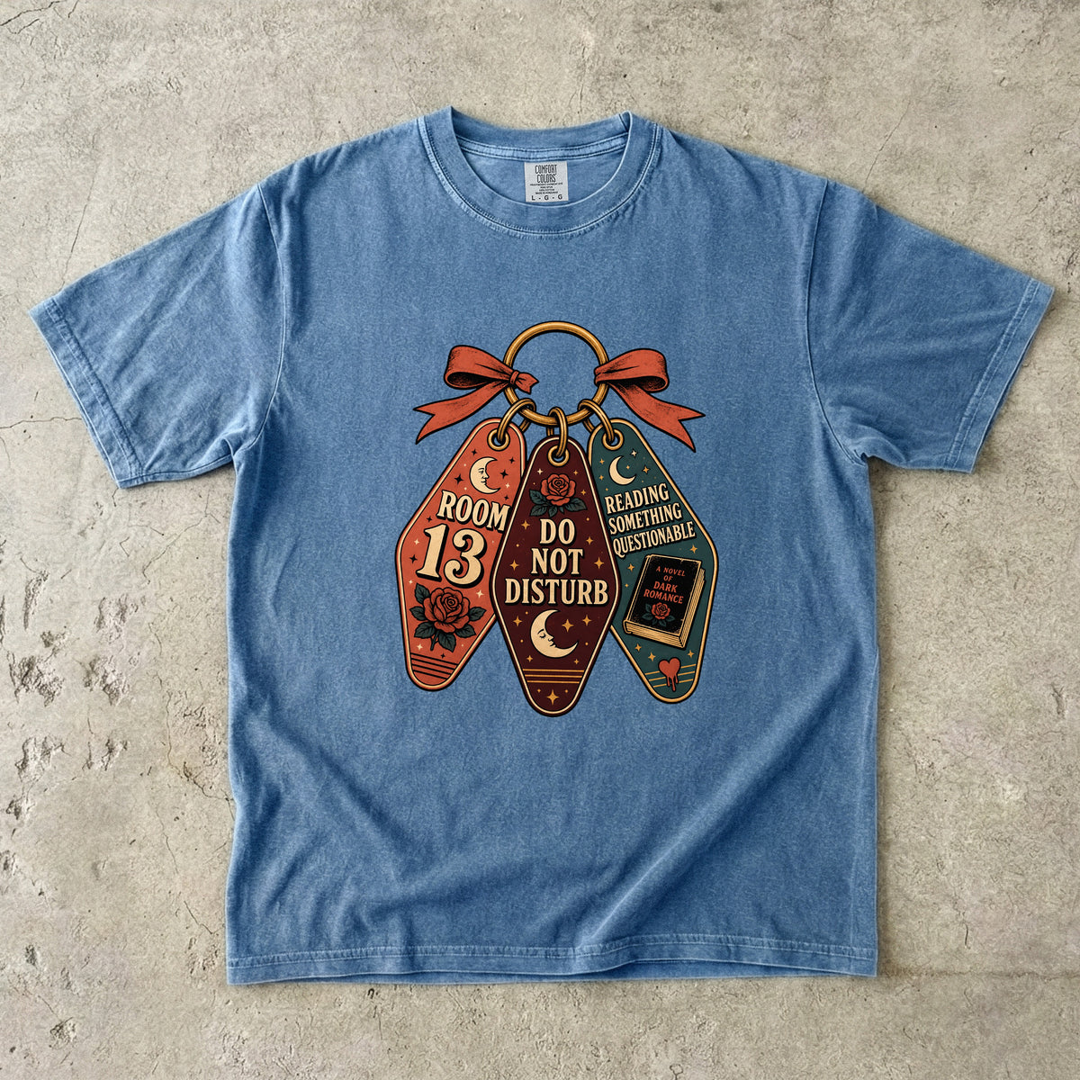 A light blue short-sleeve t-shirt laid flat on a textured concrete surface, featuring a graphic of three vintage-style library book tags tied with a red ribbon. The tags read 'ROOM 13 DO NOT DISTURB', 'READING SCHEDULE DETERMINABLE', and 'I HAVE A BOOK COMING FOR ME'.