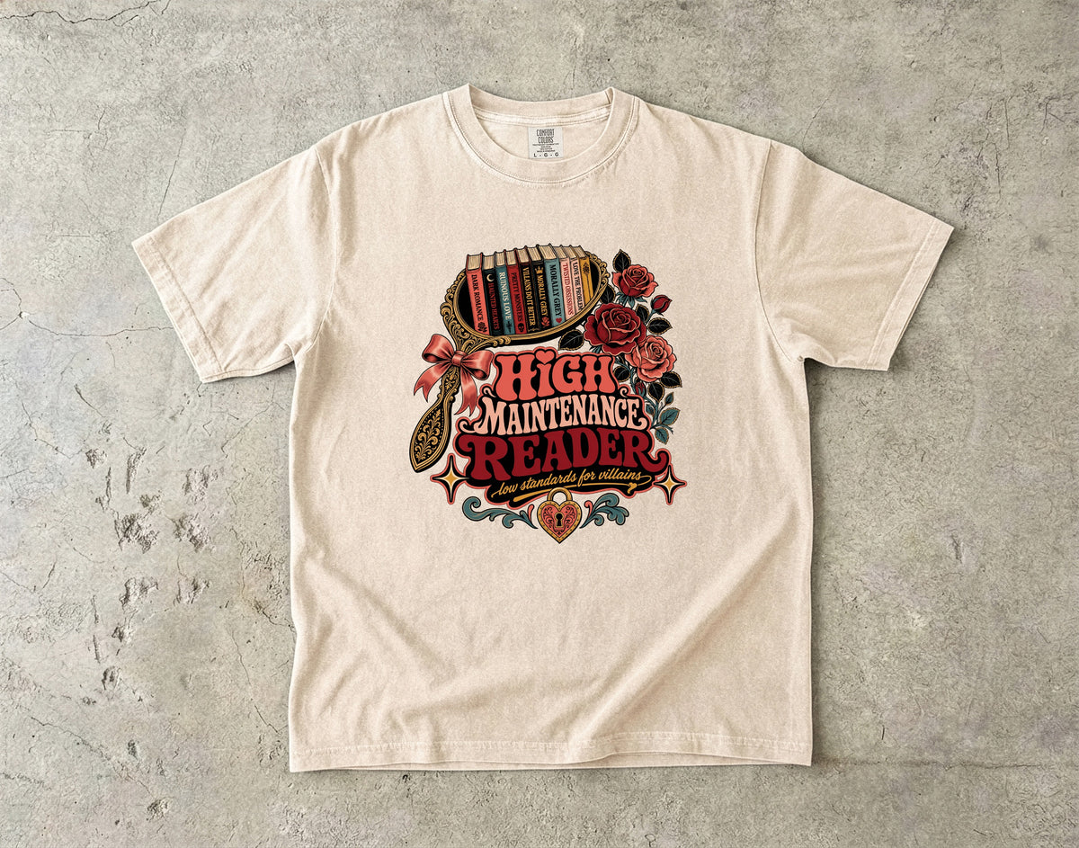An off-white crewneck t-shirt with a vintage-style graphic reading 'HIGH MAINTENANCE READER' surrounded by roses, a bookshelf, and a magnifying glass, laid flat on a textured gray concrete surface.