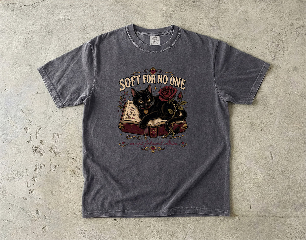 A dark gray graphic t-shirt laid flat on a textured concrete surface. The shirt features a graphic of a black cat resting on an open book next to a red rose, with the text 'SOFT FOR NO ONE' arched above the illustration.