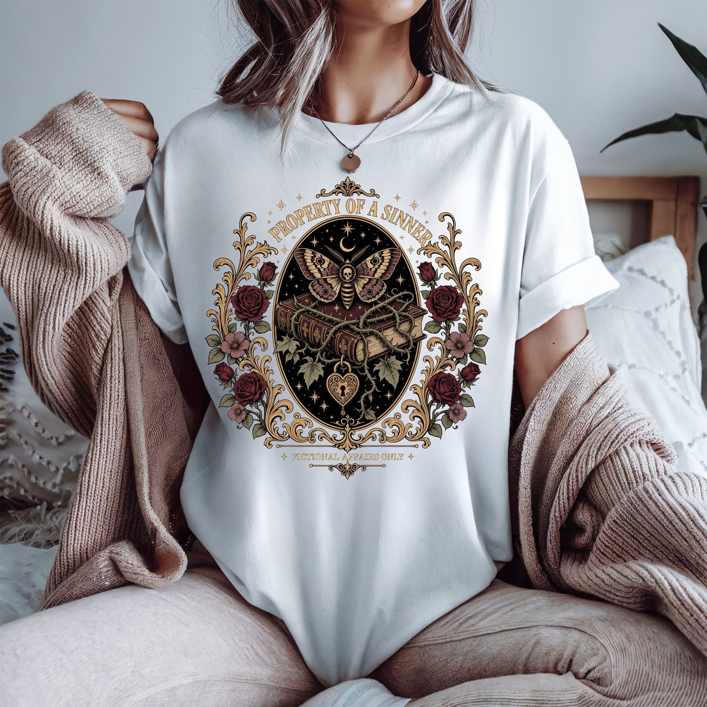 A person wearing a white oversized T-shirt with a large vintage-style butterfly and floral crest graphic on the front, styled with a beige knit sweater draped off the shoulders in a bedroom setting.