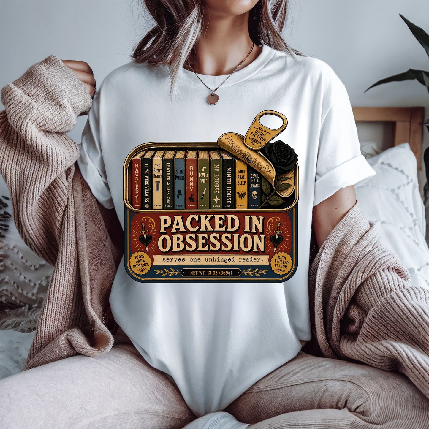 A person wearing a white graphic T-shirt sits on a bed with a beige knit sweater draped off the shoulders. The shirt features a vintage-style book tin graphic with the text 'PACKED IN OBSESSION' and illustrated books, a gold key, and a black rose.