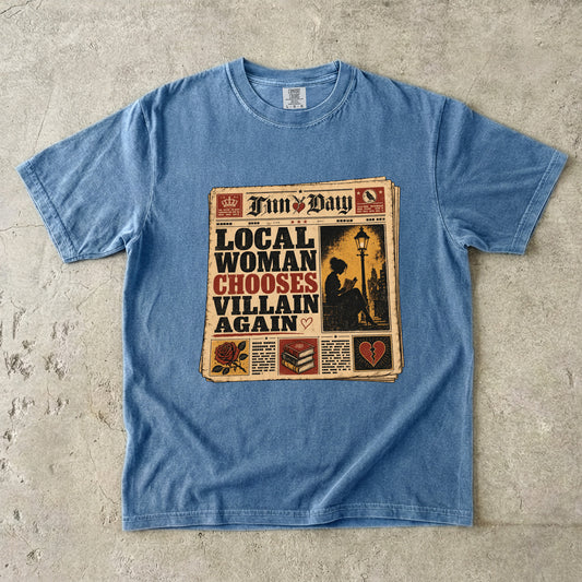 A light blue short-sleeve t-shirt laid flat on a textured concrete surface, featuring a vintage newspaper graphic on the front with the headline 'LOCAL WOMAN CHOOSES VILLAIN AGAIN' and a newspaper masthead 'Tim's Day'.