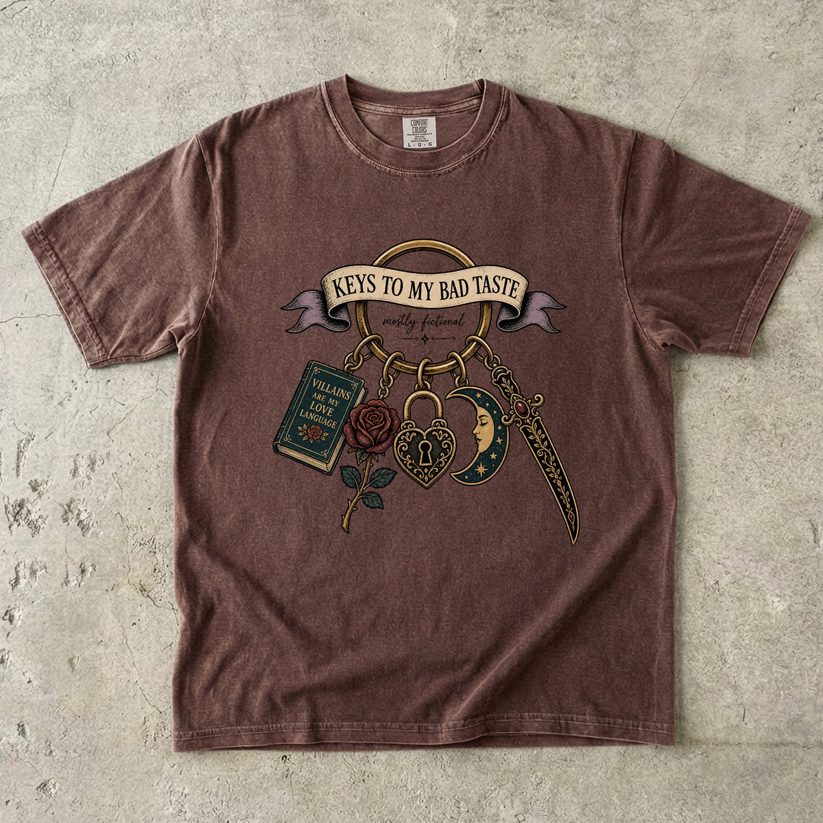 A brown short-sleeve t-shirt with a vintage-style graphic print featuring the text 'KEYS TO MY BAD TASTE' and illustrations of a book, rose, padlock, dagger, and crescent moon, laid flat on a textured concrete surface.