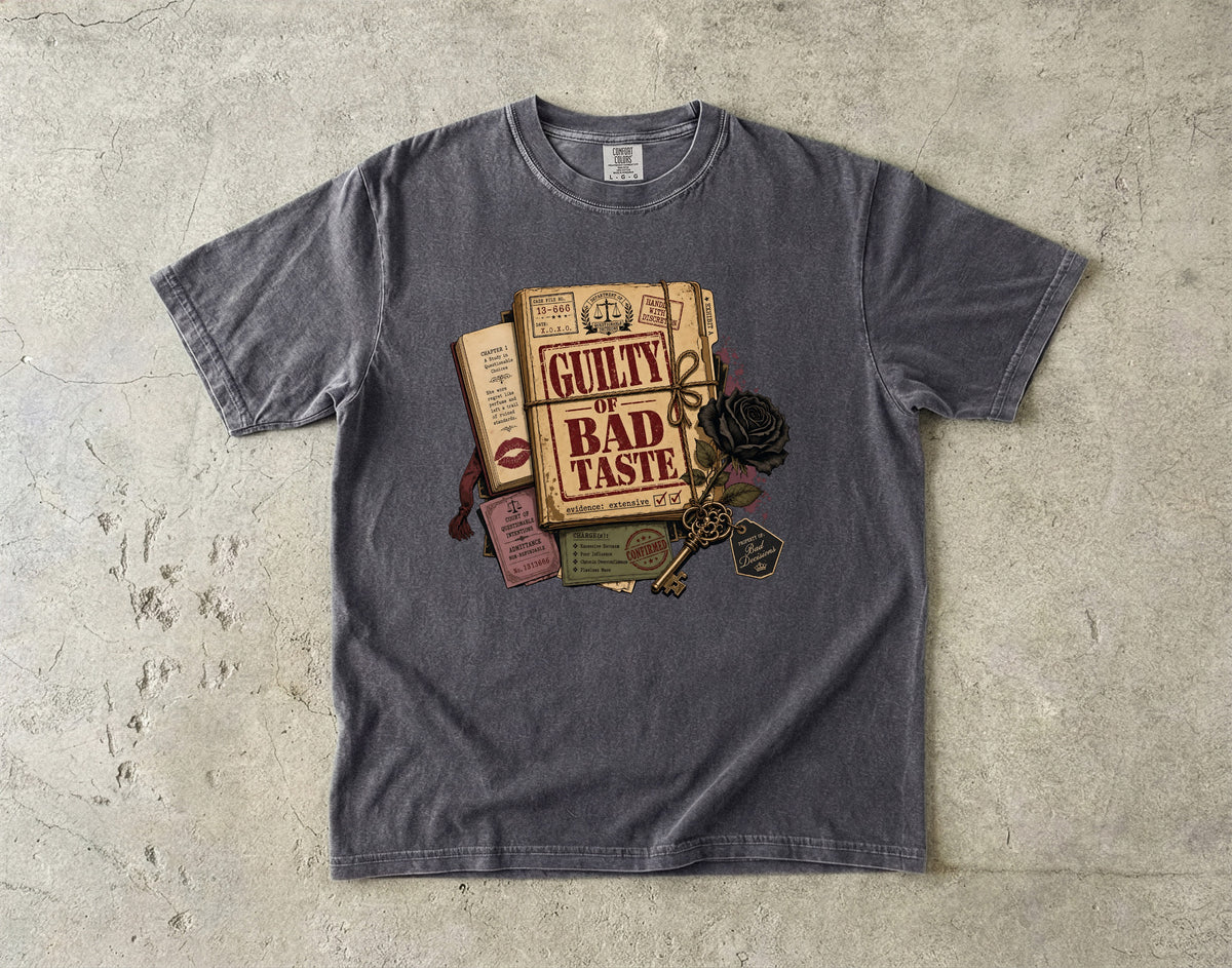 A dark gray crewneck t-shirt laid flat on a textured concrete surface, featuring a vintage-style graphic of a book titled 'GUILTY BAD TASTE' with a key, rose, and other old books.