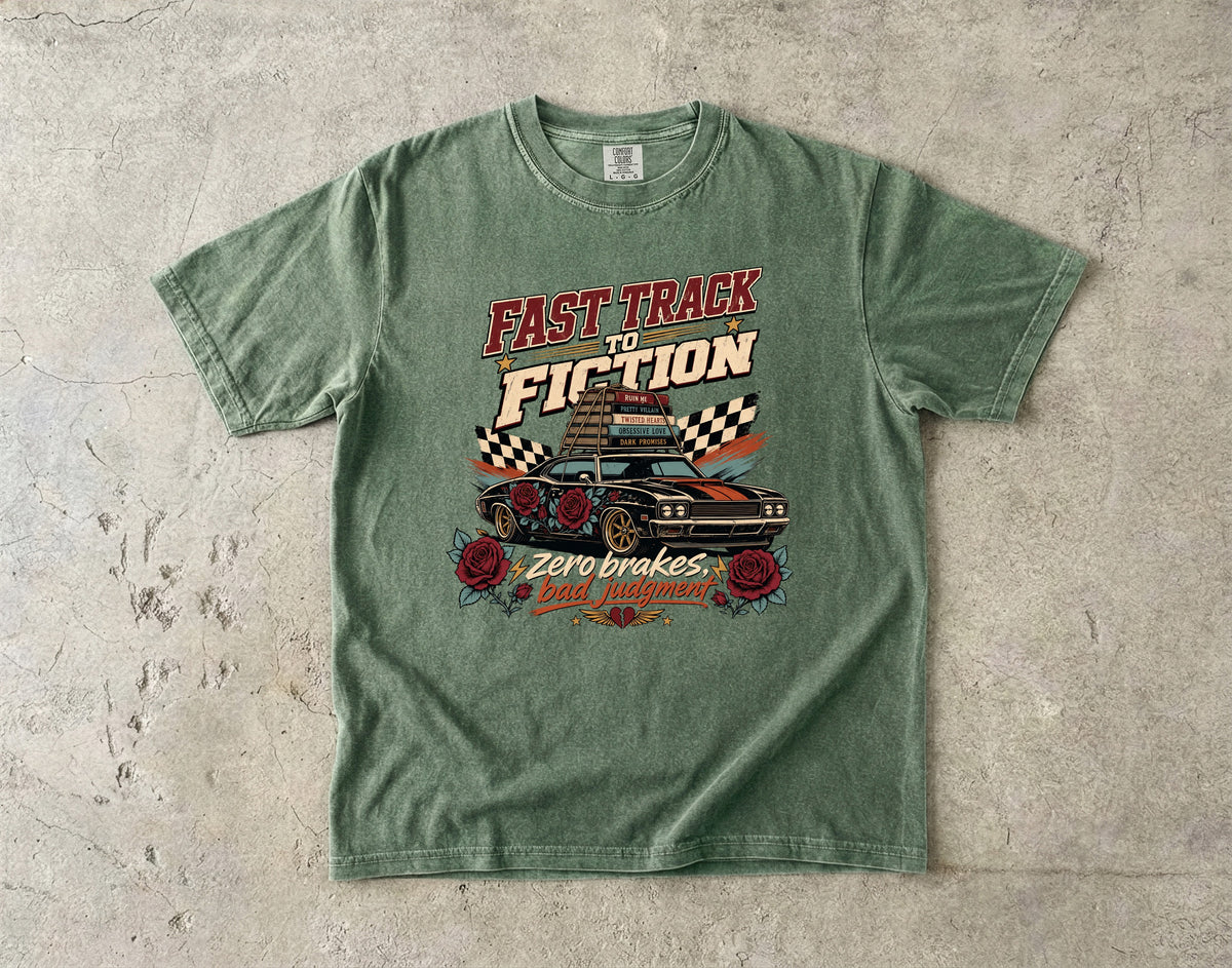 A green graphic t-shirt laid flat on a textured concrete surface. The shirt features a retro-style graphic with the text 'FAST TRACK TO FICTION' in red and cream, a black car with checkered flags, and 'Zero brakes bad attitudes' in red and cream.