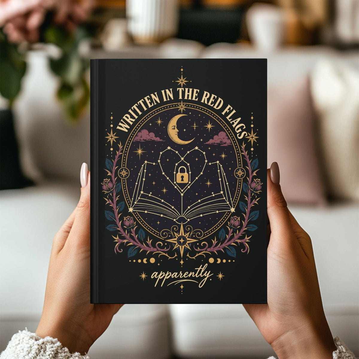 A person holds a black hardcover journal with gold and pink celestial artwork. The cover reads 'WRITTEN IN THE RED FLAGS' and 'apparently'.
