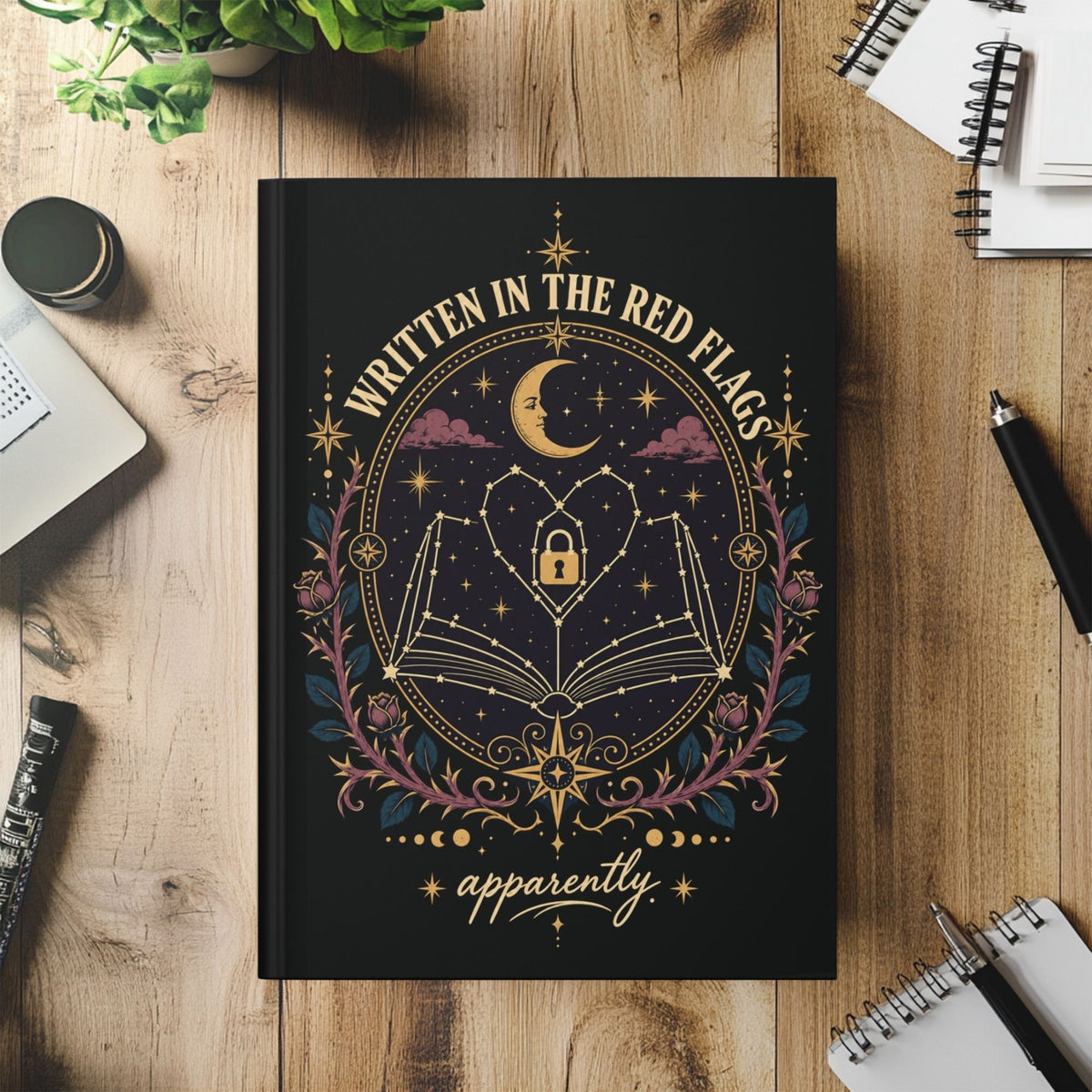 A black hardcover journal with a celestial graphic design, placed on a wooden desk surrounded by notebooks, a pen, and a small plant. The cover features a gold crescent moon, stars, an open book, a heart, and the text 'WRITTEN IN THE RED FLAGS' and 'apparently'.
