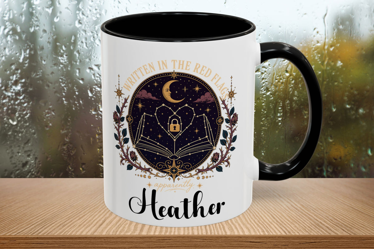 A white ceramic mug with a black handle and black interior, featuring a printed design of an open book under a crescent moon in a starry night sky, surrounded by decorative flourishes and the text 'Written in the Red Flag' and 'Heather'. The mug sits on a wooden surface with a blurred rainy window background.