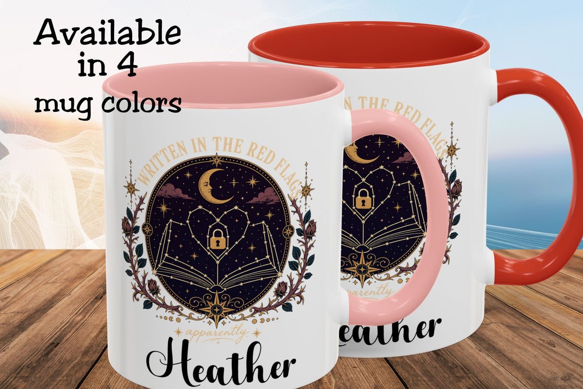 Two white ceramic mugs with pink interiors and handles are shown on a wooden surface against a blurred beach background. The mugs feature a circular celestial design with an open book, stars, a crescent moon, and the words 'Written in the Red Flag' and 'Heather.