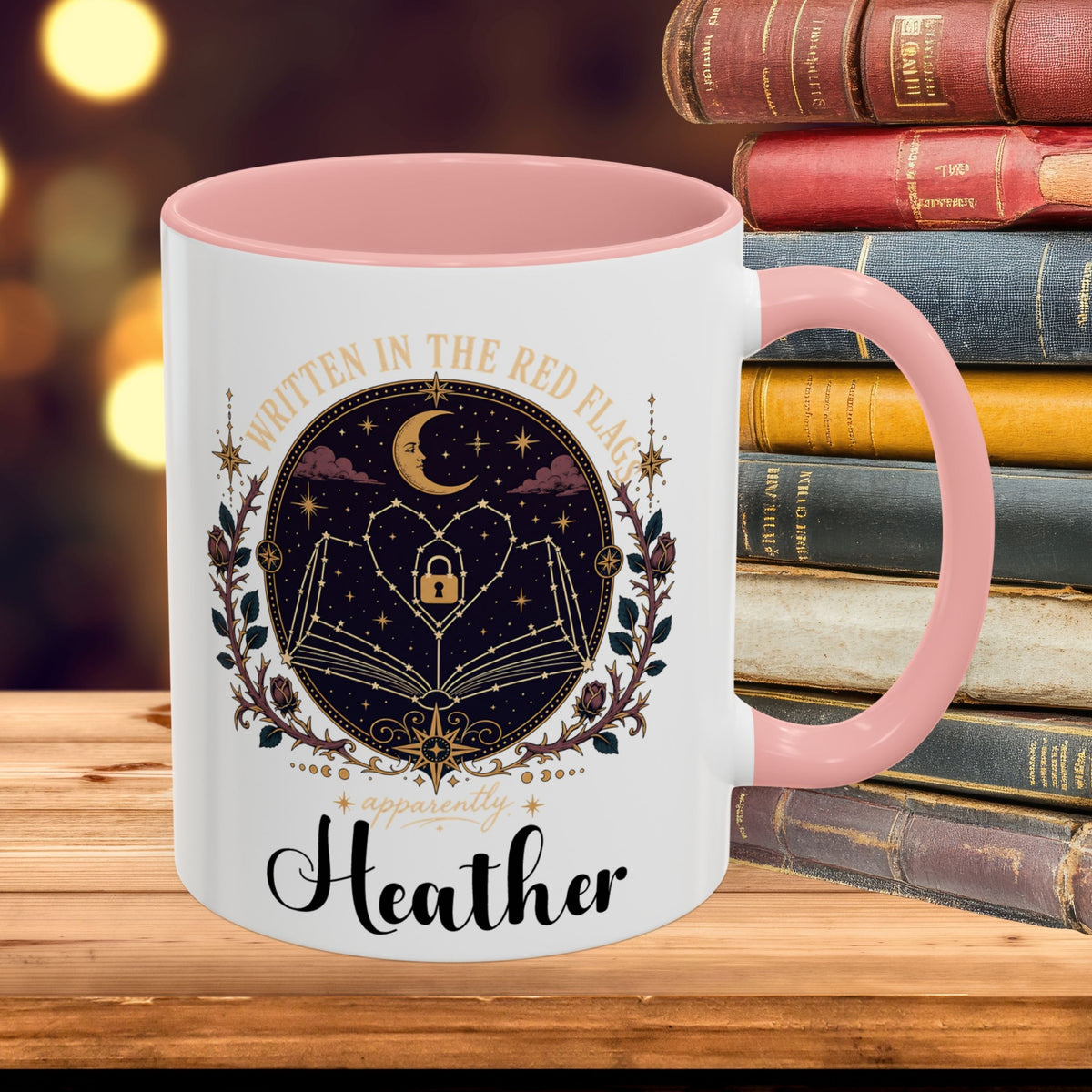 A white ceramic mug with a pink interior and handle, featuring a gothic-style graphic of an open book, stars, a crescent moon, and a lock, with the text 'Written in the Red Flag' and 'Heather'. The mug is placed on a wooden surface next to a stack of old books with a blurred warm background.