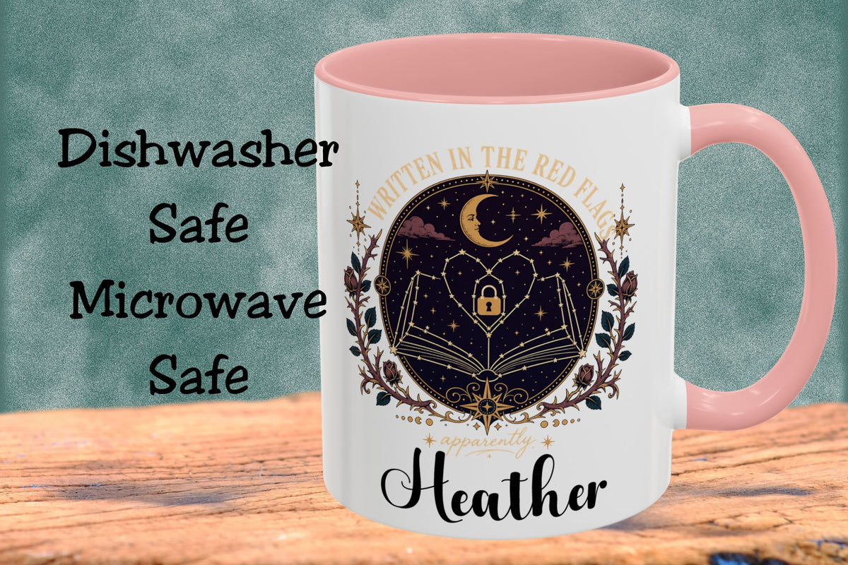 A white ceramic mug with a pink handle and interior, featuring a dark celestial graphic of a book, lock, stars, and crescent moon, with the text 'Written in the Red Flags' and 'Heather'. The mug is shown on a wooden surface against a textured teal background with text reading 'Dishwasher Safe Microwave Safe'.