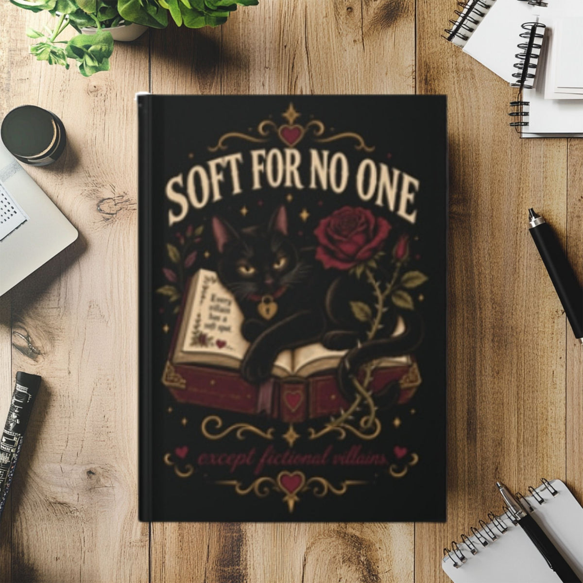 A black hardcover notebook with a gothic illustration of a black cat lounging over an open book next to a red rose, with the text 'SOFT FOR NO ONE except fictional villains' on the cover. The notebook is displayed in a flat-lay arrangement on a wooden desk surrounded by office props like pens, a laptop, and notebooks.