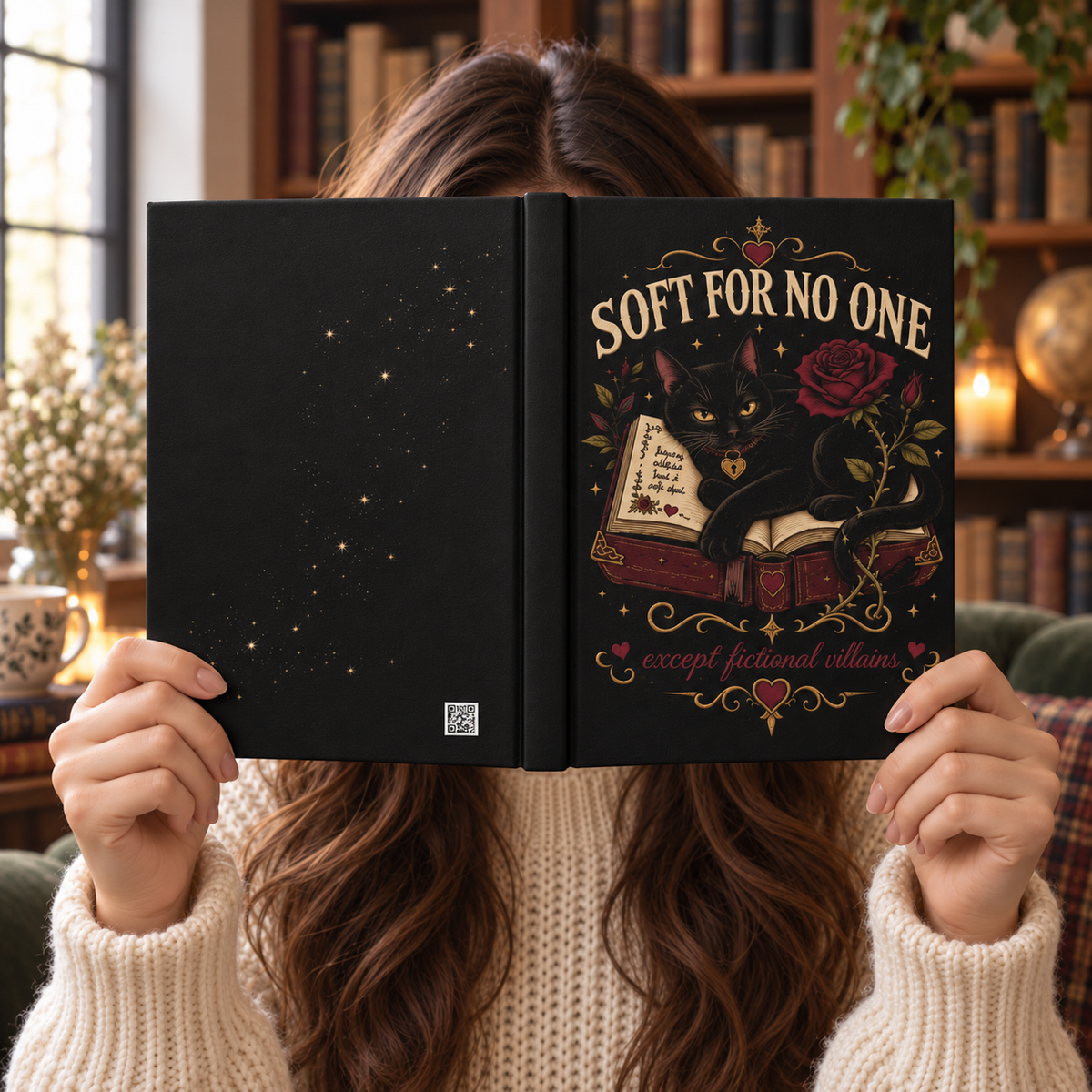 A person in a cream knit sweater holds an open hardcover book in front of their face. The book cover is black with gold decorative text reading 'SOFT FOR NO ONE' and an illustration of a black cat on an open book with a red rose.