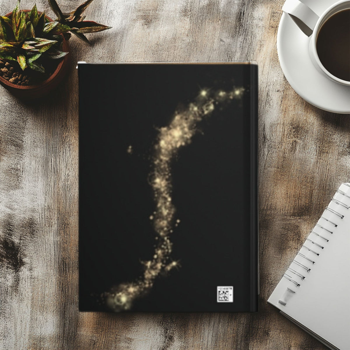A black hardcover notebook with a glowing gold cosmic dust trail design on the cover, styled in a flat-lay on a rustic wooden table next to a cup of coffee, a small potted succulent, and a spiral notebook.