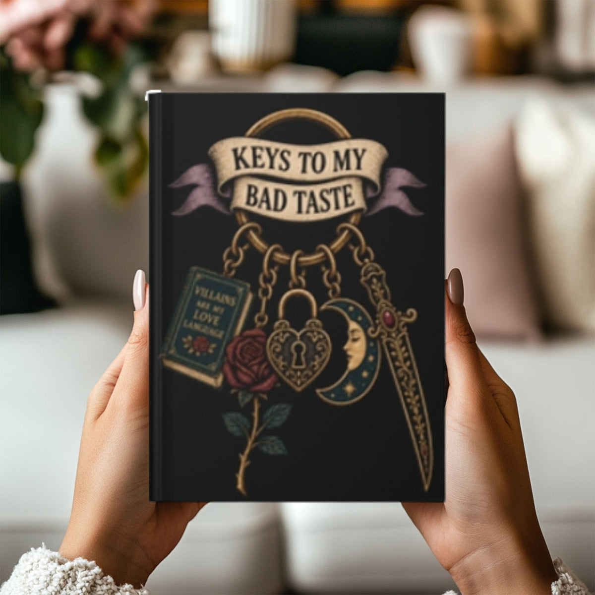 A person holds a black hardcover journal with a gothic illustration that reads 'KEYS TO MY BAD TASTE' above a rose, padlock, dagger, and book. The background is a softly blurred living room interior.