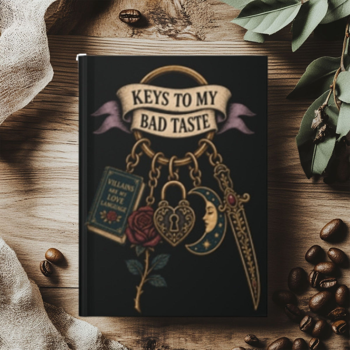 A black hardcover notebook with gothic-themed keychain illustrations, including a rose, padlock, sword, book, and crescent moon, with the text 'KEYS TO MY BAD TASTE' on the cover. The notebook is styled on a rustic wooden surface with coffee beans, a linen cloth, and green leaves.