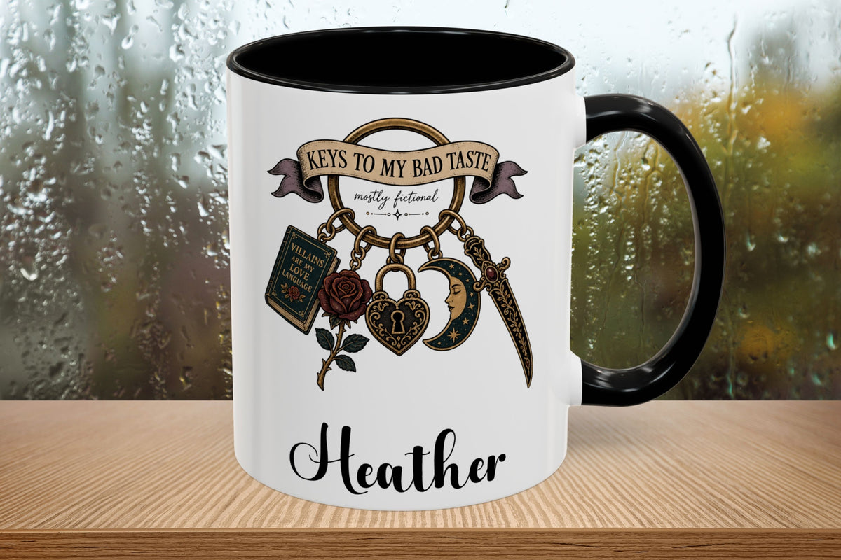 A white ceramic mug with a black handle and interior, featuring a gothic-style graphic with the text 'Keys to My Bad Taste' and the name 'Heather' printed on the front. The mug is placed on a wooden surface with a blurred, rainy window background.
