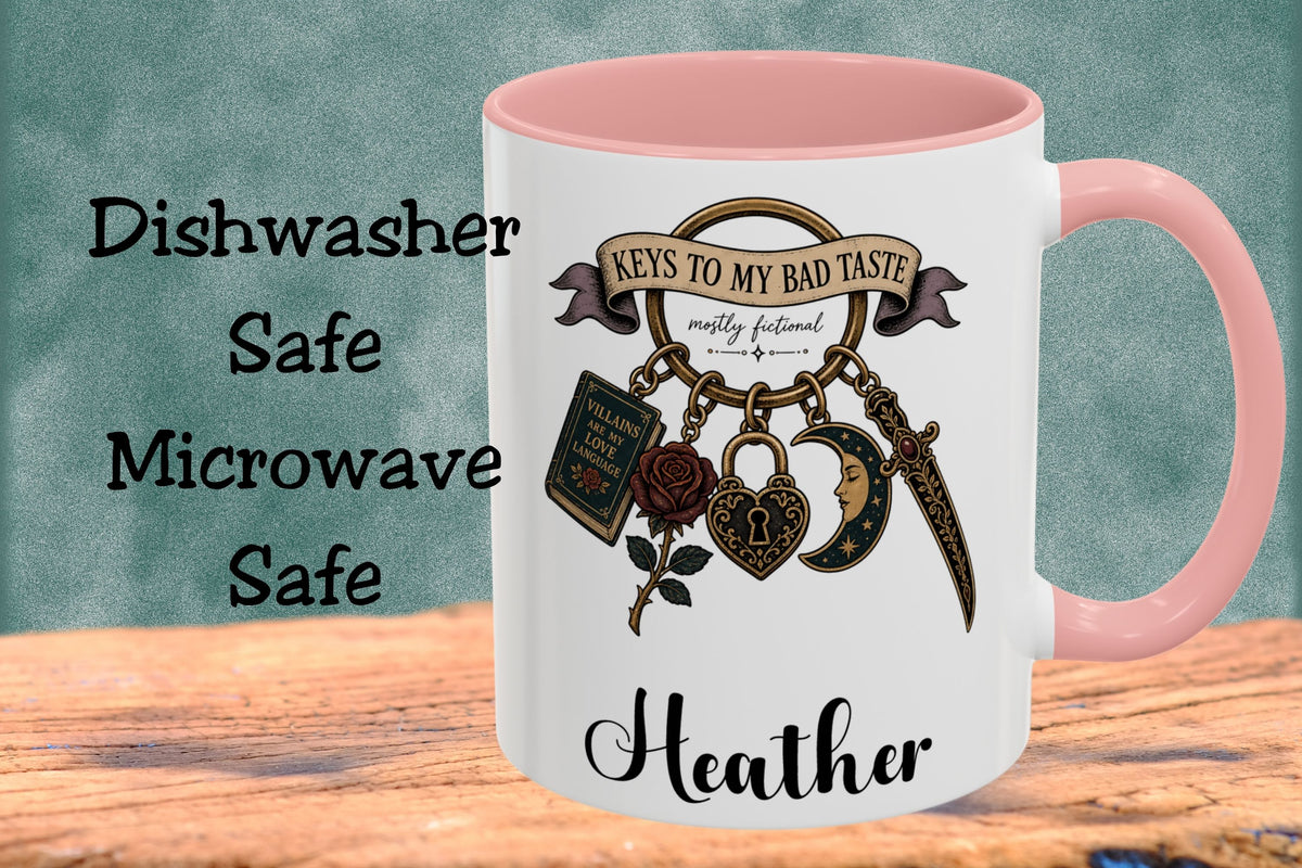 A white ceramic mug with a pink interior and handle, featuring a graphic of vintage-style keys and a banner that reads 'KEYS TO MY BAD TASTE' and 'mostly fictional.' The mug is shown on a wooden surface against a textured teal background, with text on the left reading 'Dishwasher Safe Microwave Safe' and the name 'Heather' printed on the mug.