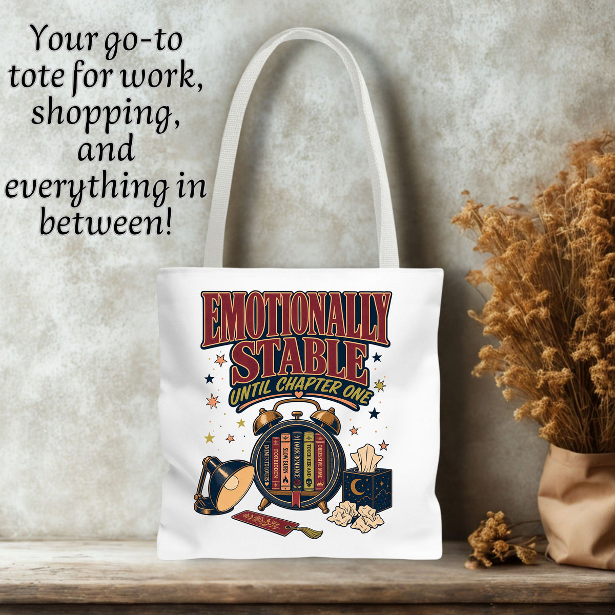 A white canvas tote bag with a colorful graphic print reading 'EMOTIONALLY STABLE UNTIL CHAPTER ONE' and featuring a bookshelf, lamp, and popcorn bucket, displayed on a wooden surface against a textured gray wall with dried flowers and text overlay.