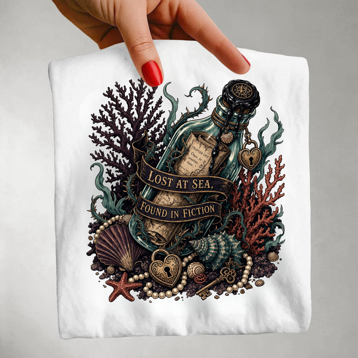 A hand with red nail polish holds up a white t-shirt featuring a detailed graphic of a 'Lost at Sea, Found in Fiction' treasure map bottle surrounded by seashells, coral, pearls, and a heart-shaped lock.