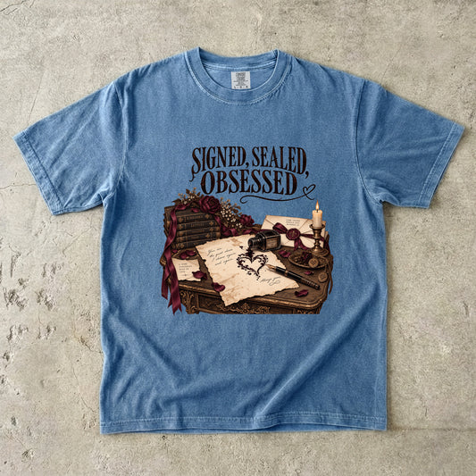 A light blue crewneck t-shirt laid flat on a textured concrete surface. The shirt features a graphic print of a vintage desk with a quill, inkwell, candle, and letter, topped with the gothic text 'SIGNED, SEALED, OBSESSED'.