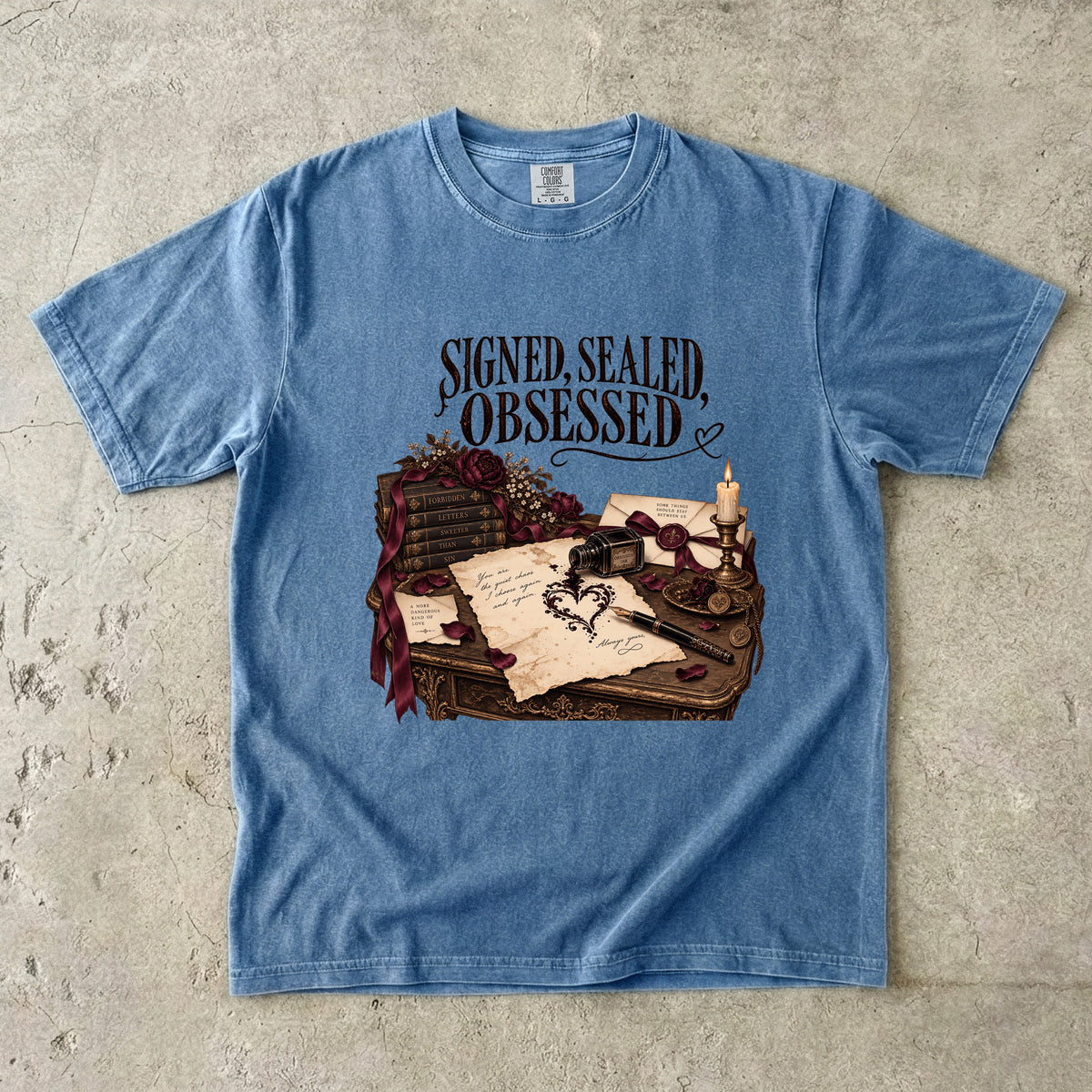 A light blue crewneck t-shirt laid flat on a textured concrete surface. The shirt features a graphic print of a vintage desk with a quill, inkwell, candle, and letter, topped with the gothic text 'SIGNED, SEALED, OBSESSED'.