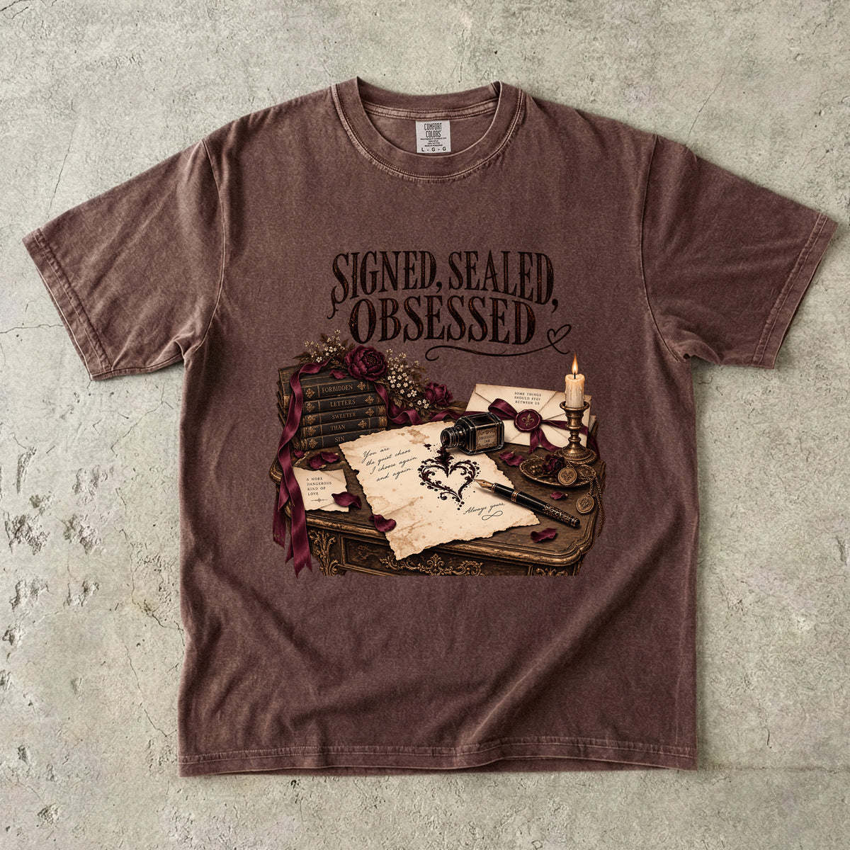 A brown short-sleeve graphic t-shirt laid flat on a textured gray concrete background. The shirt features a vintage-style illustration of a desk with an inkwell, quill, candle, and open book, with the text 'SIGNED, SEALED, OBSESSED' printed above the graphic.