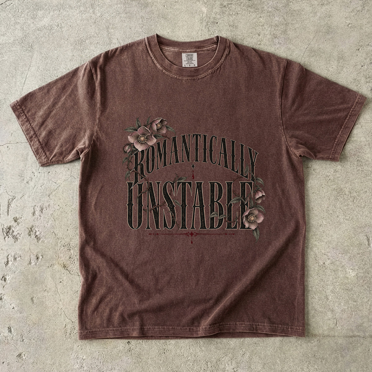 A brown short-sleeve t-shirt with a distressed acid-wash finish, featuring a graphic print that reads 'ROMANTICALLY UNSTABLE' in dark green letters with pink roses and green leaves on a textured concrete background.
