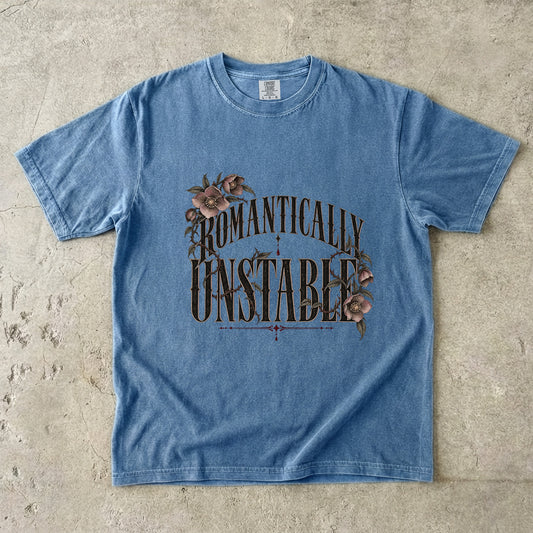A light blue short-sleeve t-shirt with a distressed graphic reading 'ROMANTICALLY UNSTABLE' in dark brown lettering, decorated with pink roses, laid flat on a textured concrete surface.