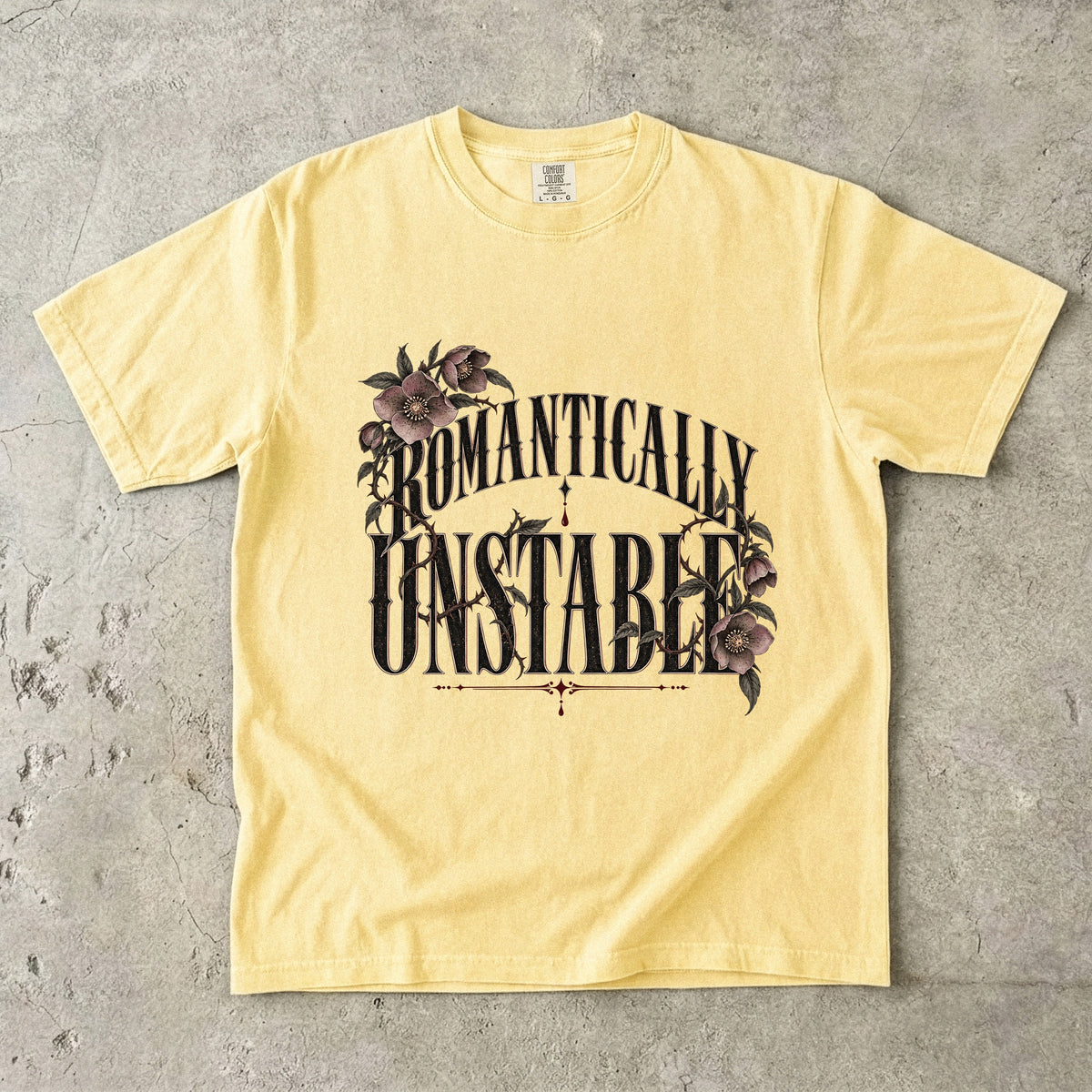 A pale yellow short-sleeve t-shirt with a graphic print reading 'ROMANTICALLY UNSTABLE' in black stylized lettering, decorated with dark purple flowers and green leaves, laid flat on a textured gray concrete surface.