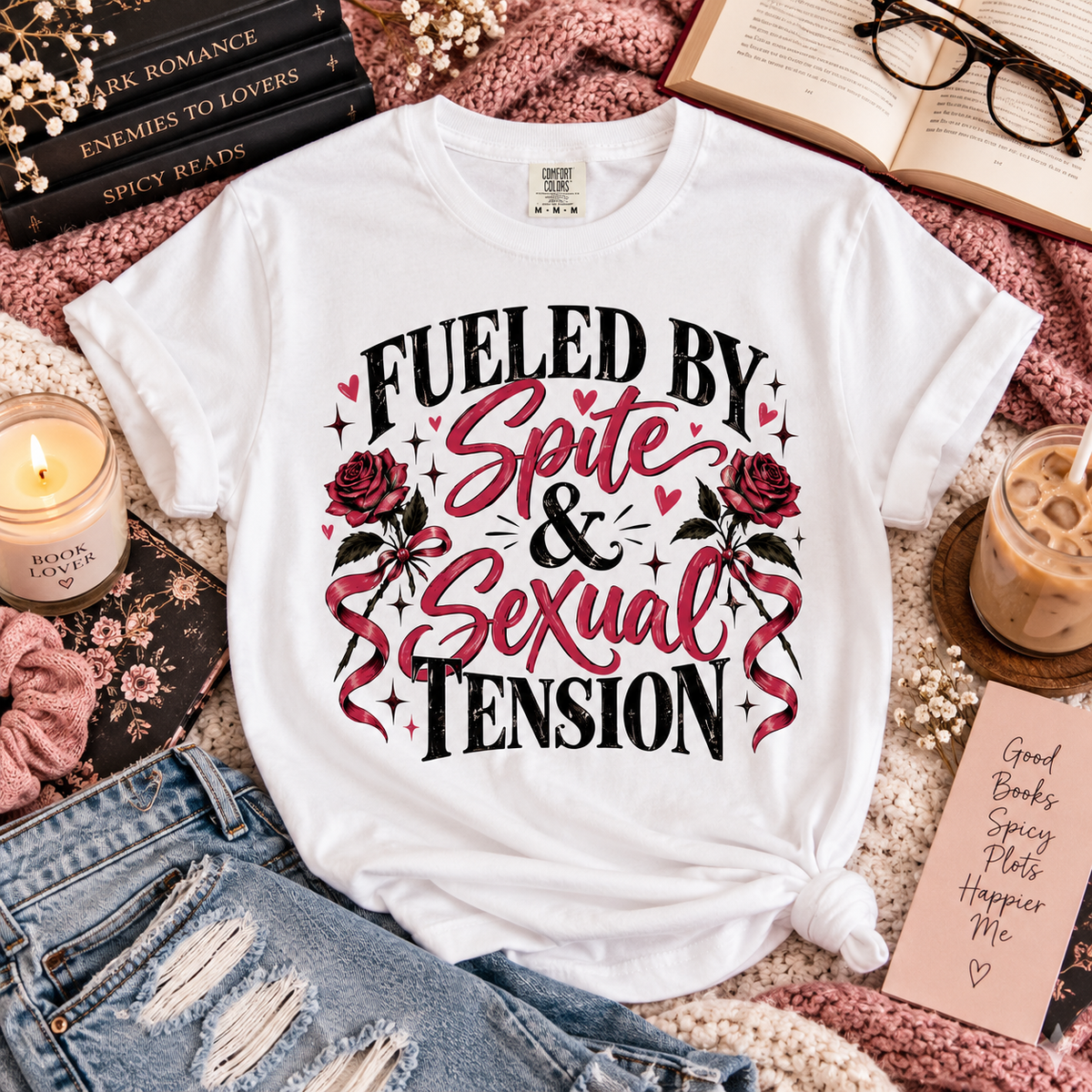 A white graphic T-shirt laid flat in a styled arrangement with denim shorts, an open book, glasses, a candle, an iced coffee, and a note card. The shirt has a large front print reading “FUELED BY Spite & Sexual Tension” in black and pink lettering with roses and sparkles.