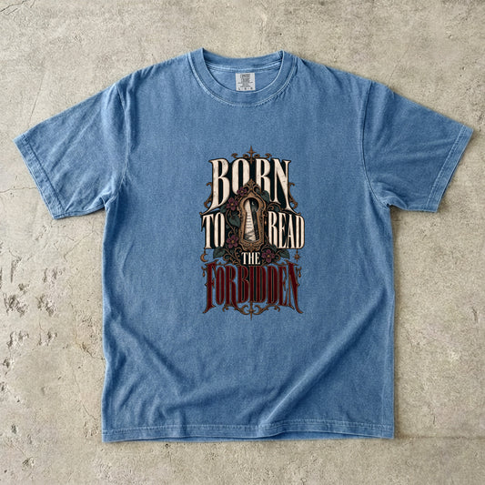 A light blue short-sleeve graphic t-shirt laid flat on a textured concrete surface. The graphic features the text 'BORN TO READ THE FORBIDDEN' in a stylized, vintage font with a dark, ornate illustration in the center.