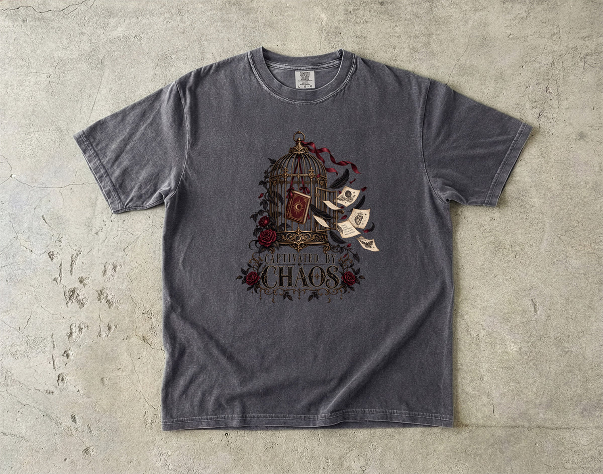 A dark gray crewneck t-shirt with a graphic print of a birdcage, red roses, flying parchment scrolls, and the text 'BURN THE CHAOS' laid flat on a textured concrete surface.