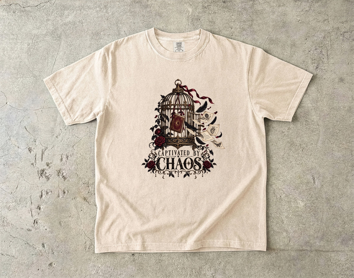 A cream-colored short-sleeve t-shirt with a dark graphic print of a birdcage, roses, and the words 'Seduced by Chaos' laid flat on a textured gray concrete surface.