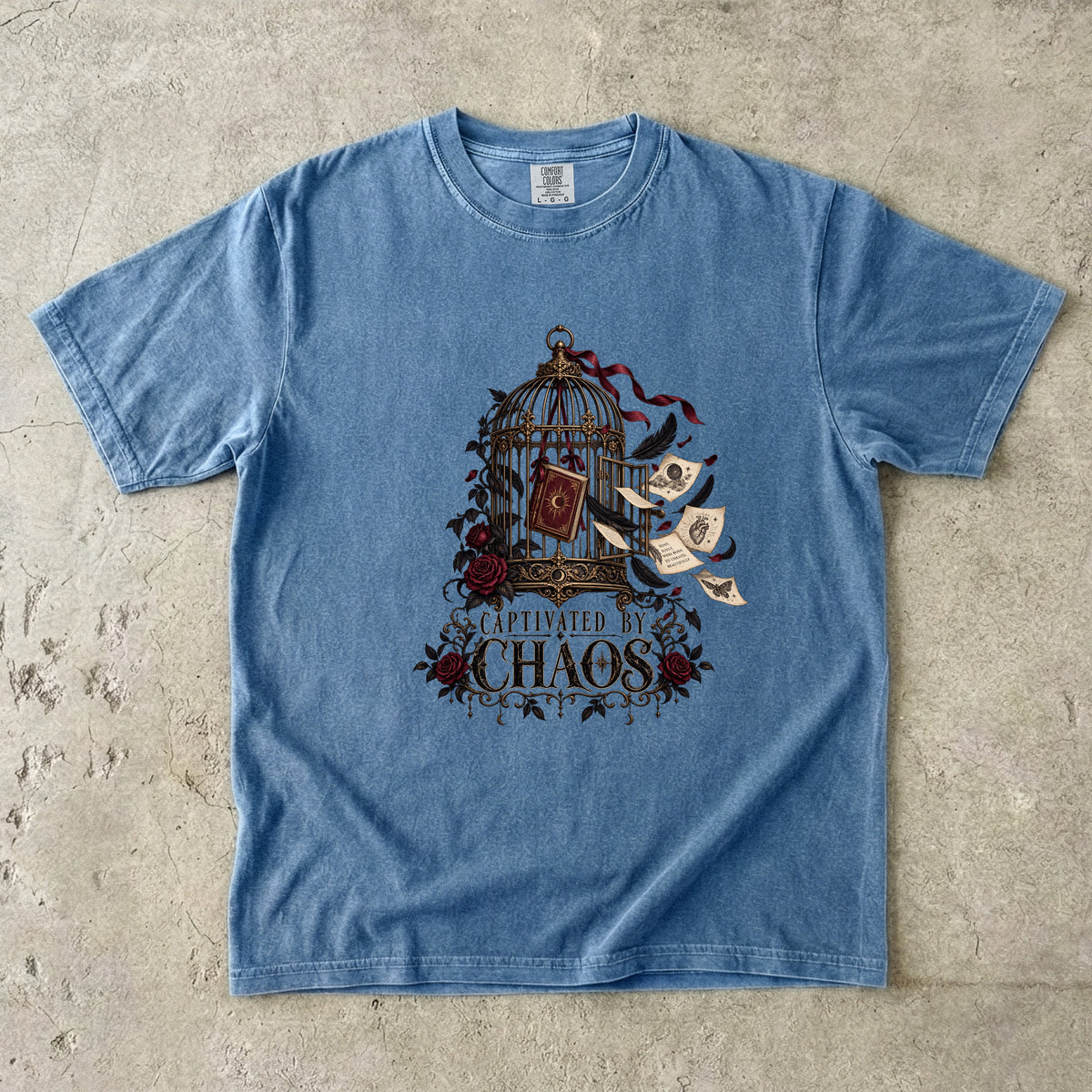 A light blue short-sleeve t-shirt with a vintage-style graphic on the front featuring a birdcage, flying scrolls, roses, and the text 'CAPTIVATED IN CHAOS'. The shirt is laid flat on a textured concrete surface.