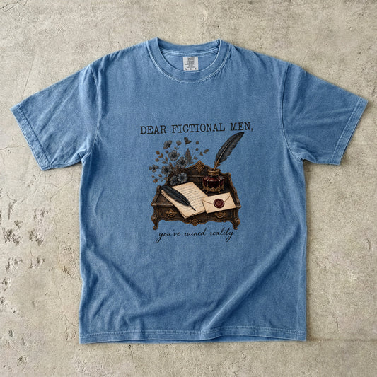 A light blue graphic t-shirt laid flat on a textured concrete surface. The shirt features a vintage-style illustration of an inkwell, quill, and open book on a desk, with the text 'DEAR FICTIONAL MEN,' above the graphic and 'you're meant really' below.