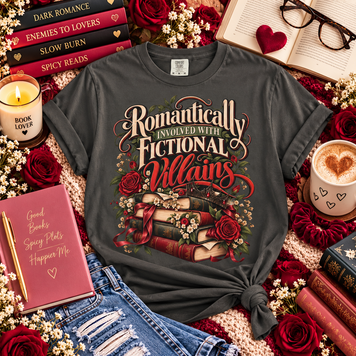 A dark gray graphic t-shirt with a romantic book-themed design, styled in a cozy flat-lay arrangement with books, roses, a candle, and a coffee cup.