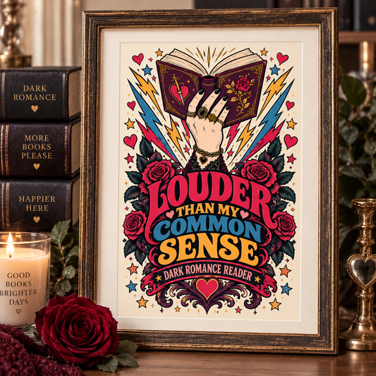 A framed art print featuring a hand holding an open book surrounded by roses, stars, and lightning, with the text 'LOUDER THAN MY COMMON SENSE' and 'DARK ROMANCE Reader'. The frame is styled on a wooden surface next to a candle, a red rose, and stacked books.