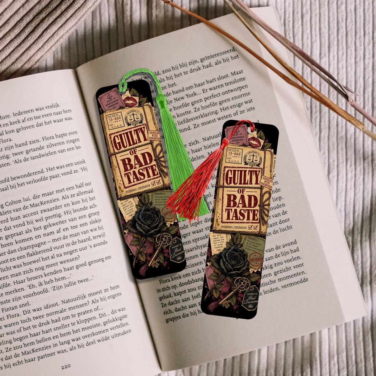 Two decorative bookmarks shaped like whiskey glasses with 'GUILTY BAD TASTE' labels, displayed on an open book. One bookmark has a green tassel and the other has a red tassel, with wooden chopsticks resting on the right side.