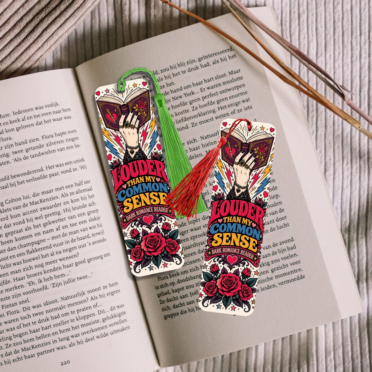 Two decorative bookmarks with a colorful tattoo-style illustration of a woman and roses, placed on an open book with two wooden sticks on a light wood surface.