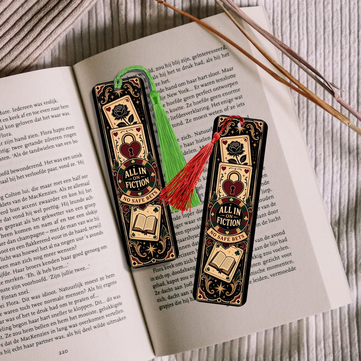 Two decorative book bookmarks styled on an open book, featuring a black and gold design with a central red heart and the words 'ALL IN FICTION'. One bookmark has a green tassel and the other has a red tassel, with wooden chopsticks resting on the right side.