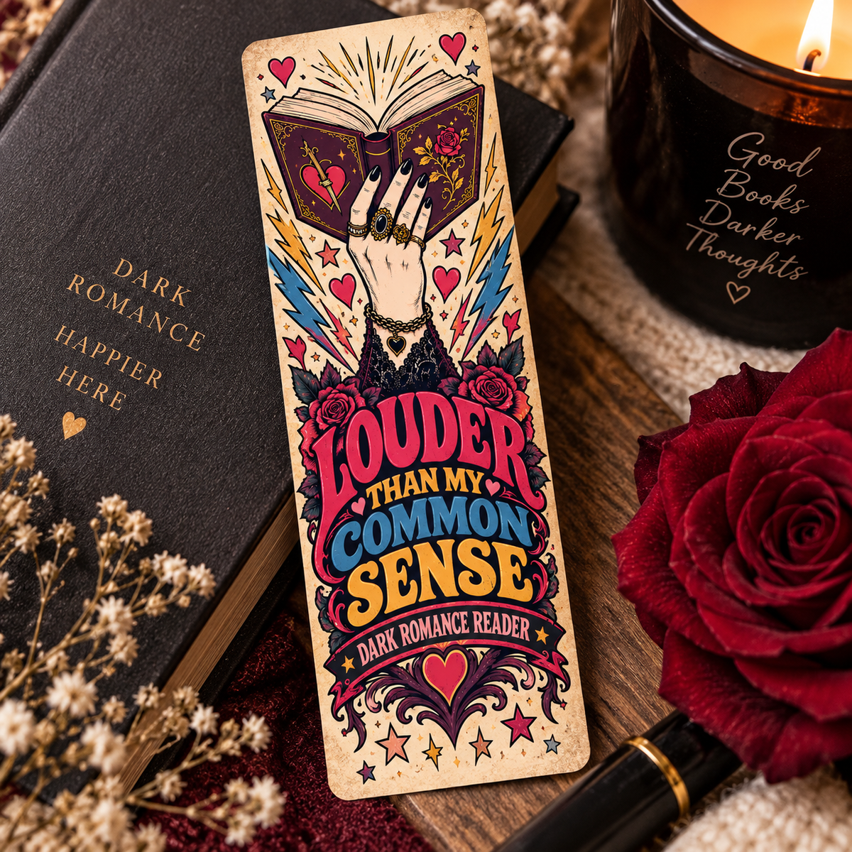 A decorative rectangular book bookmark or card featuring an illustrated hand holding an open book with the text 'Louder than my common sense' and 'Dark Romance Reader' at the bottom, styled on a wooden surface with a black book, candle, rose, and dried flowers.