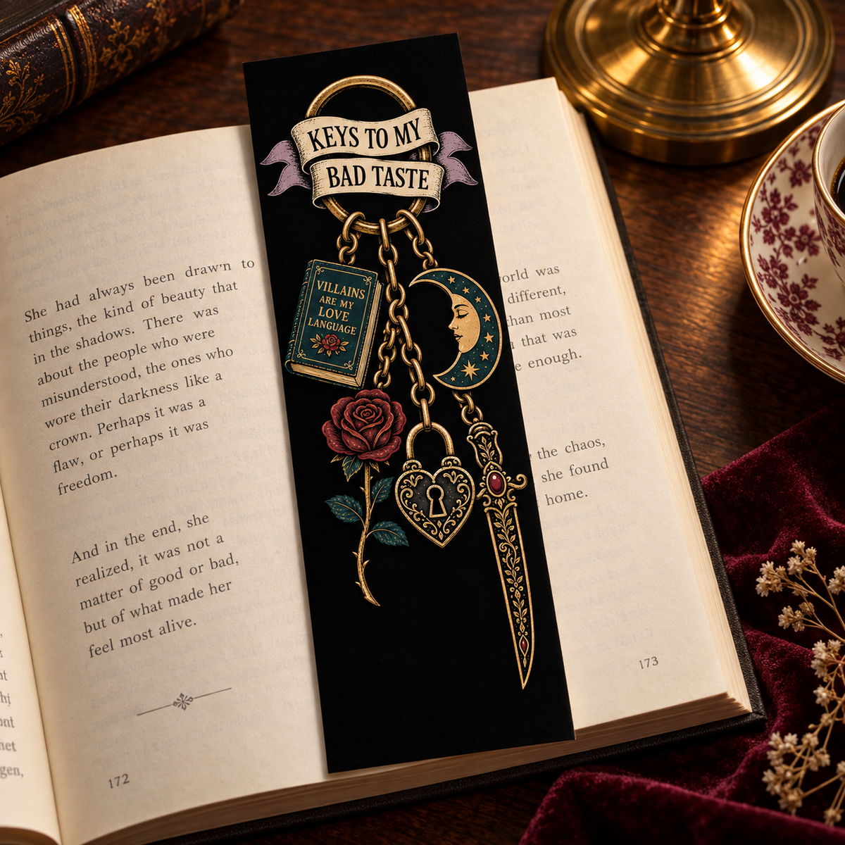 A decorative black bookmark styled on an open book, featuring a gold-toned chain design with a crescent moon, heart, rose, and a banner reading 'KEYS TO MY BAD TASTE'. The scene is set on a dark wooden table with a brass lamp base, a cup, and a leather-bound book in the background.