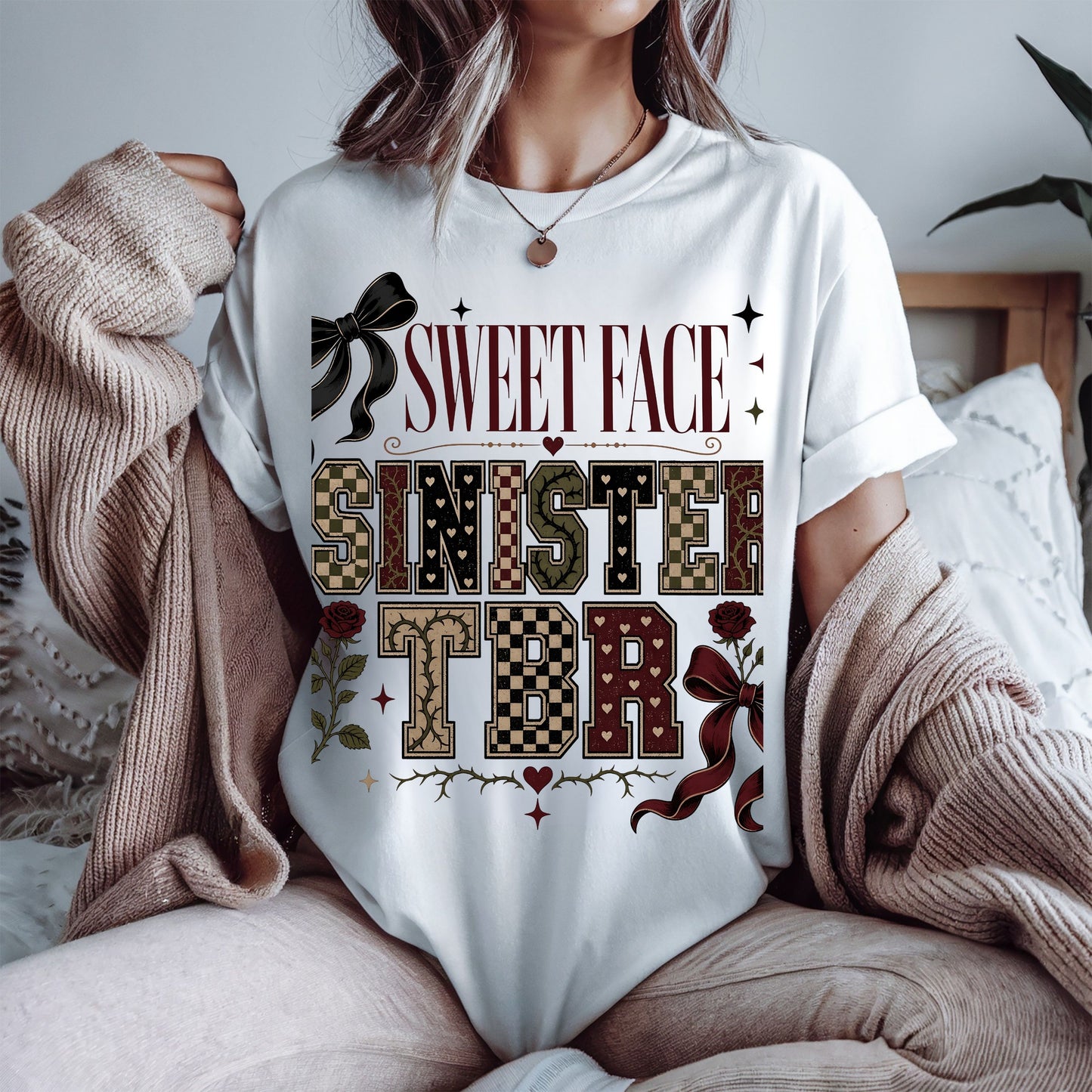 A woman wearing a white graphic t-shirt with a 'SWEET FACE SINISTERS TBR' design in a vintage western theme, styled with a beige knit cardigan in a cozy bedroom setting.