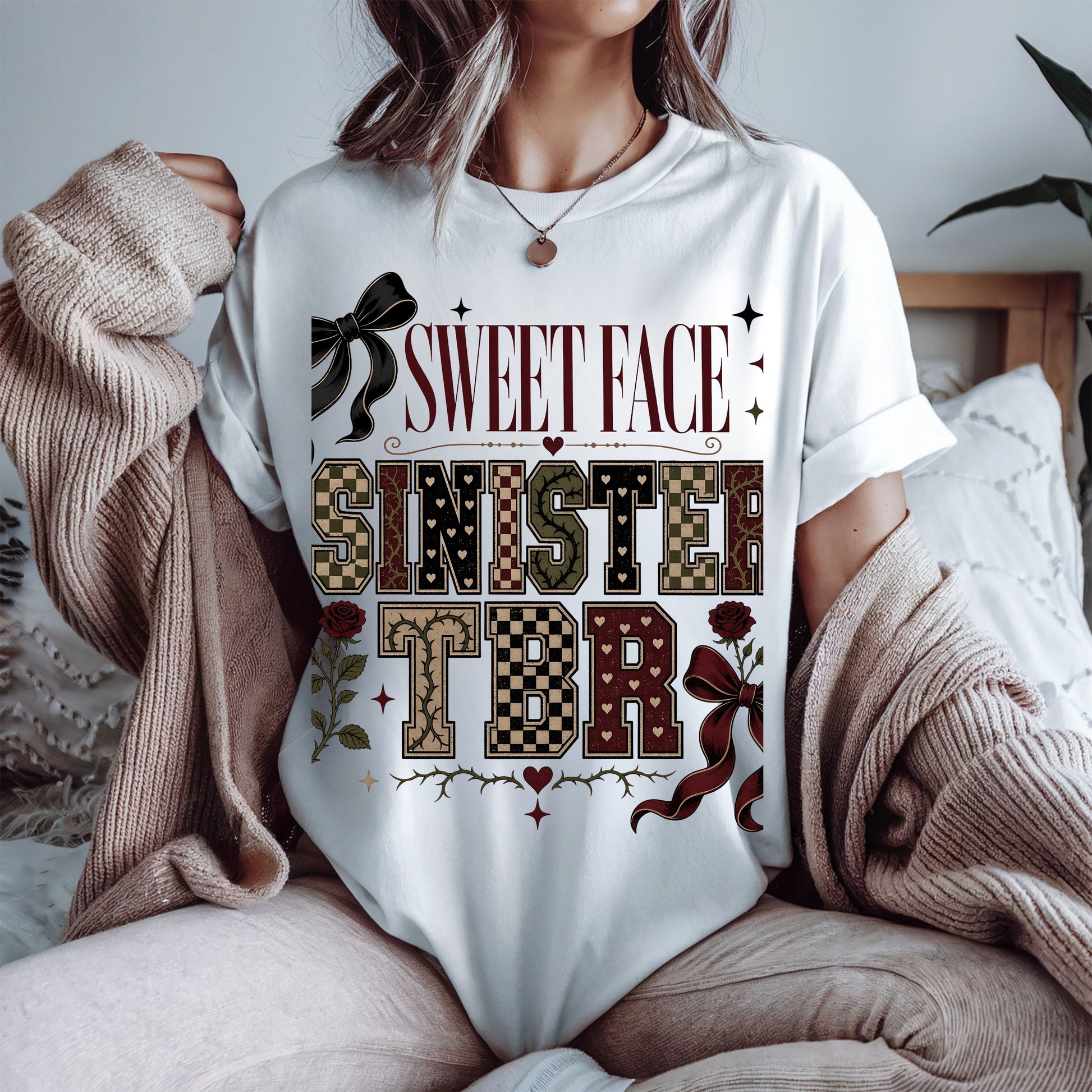 A woman wearing a white graphic t-shirt with a 'SWEET FACE SINISTERS TBR' design in a vintage western theme, styled with a beige knit cardigan in a cozy bedroom setting.