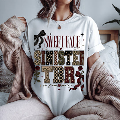 A woman wearing a white graphic t-shirt with a 'SWEET FACE SINISTERS TBR' design in a vintage western theme, styled with a beige knit cardigan in a cozy bedroom setting.