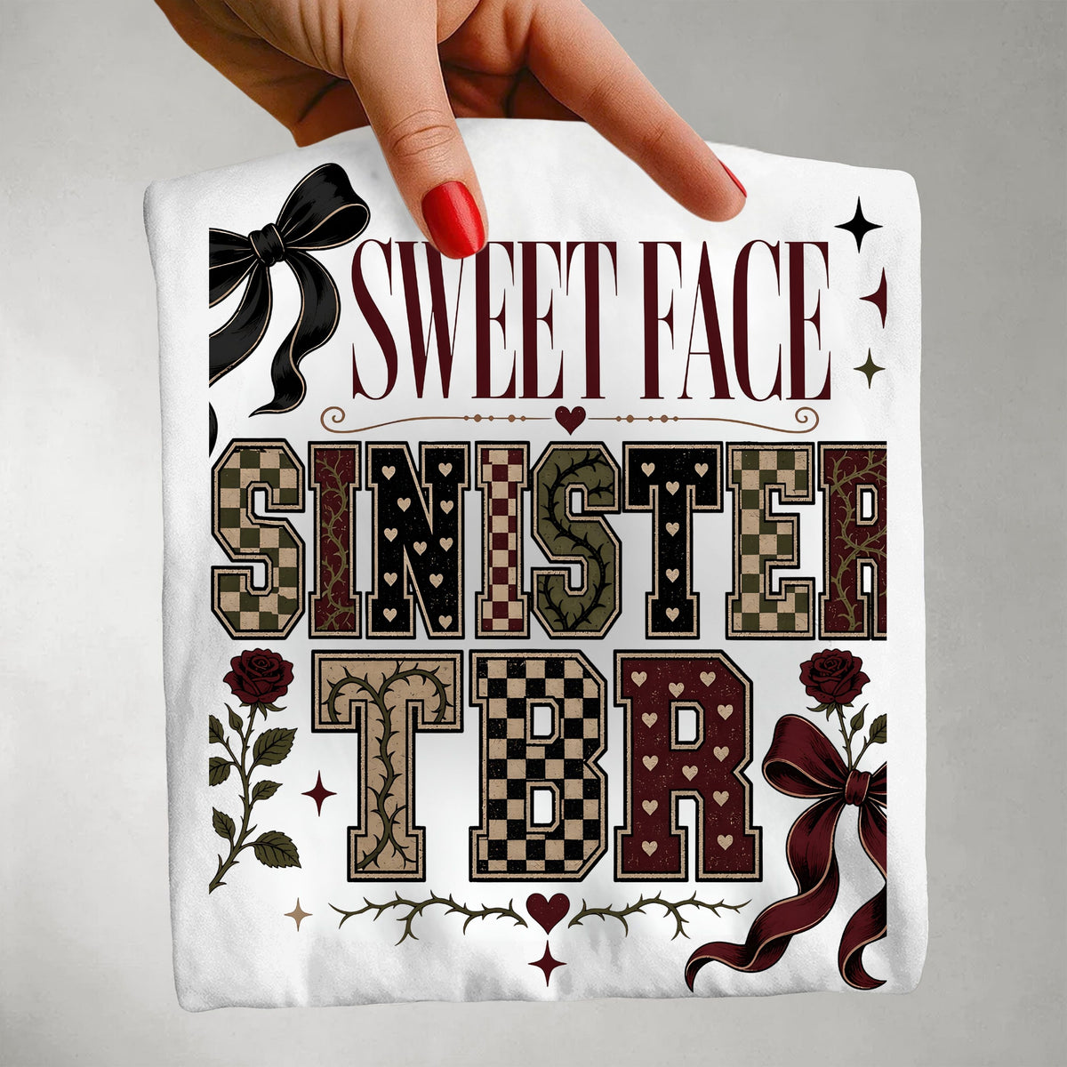 A hand with red nail polish holds a white square fabric item, likely a napkin or small cloth, printed with the phrase 'SWEET FACE SINISTER TBR' in decorative lettering with bows, roses, stars, and thorny accents on a light gray background.