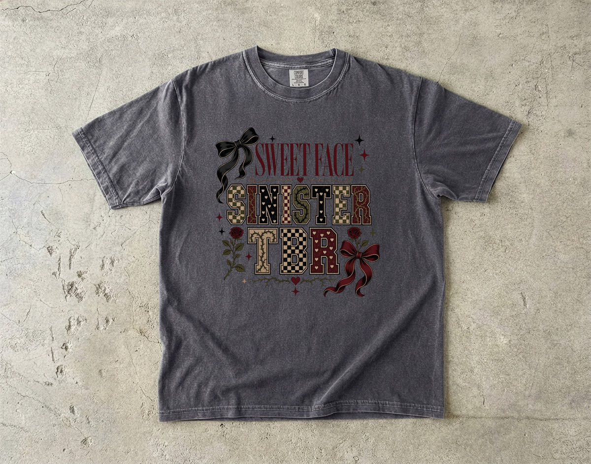 A dark gray graphic t-shirt laid flat on a textured concrete surface. The shirt features a festive graphic that reads 'SWEET FACE SINISTER' in red, black, and patterned lettering, decorated with red bows and small red hearts.