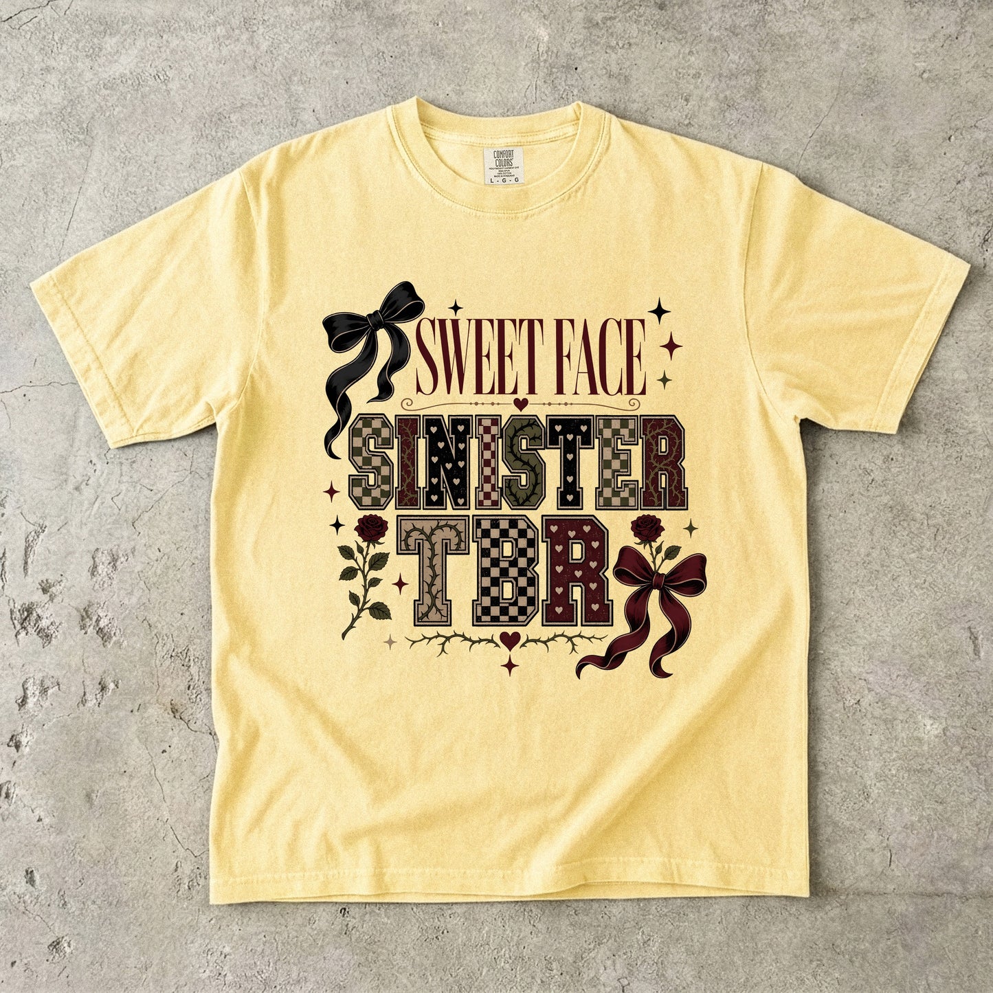 A pale yellow graphic t-shirt laid flat on a textured gray concrete background. The shirt features a gothic-style graphic with the words 'SWEET FACE SINISTER TDR' in a patterned font, decorated with black and dark red bows, roses, and stars.
