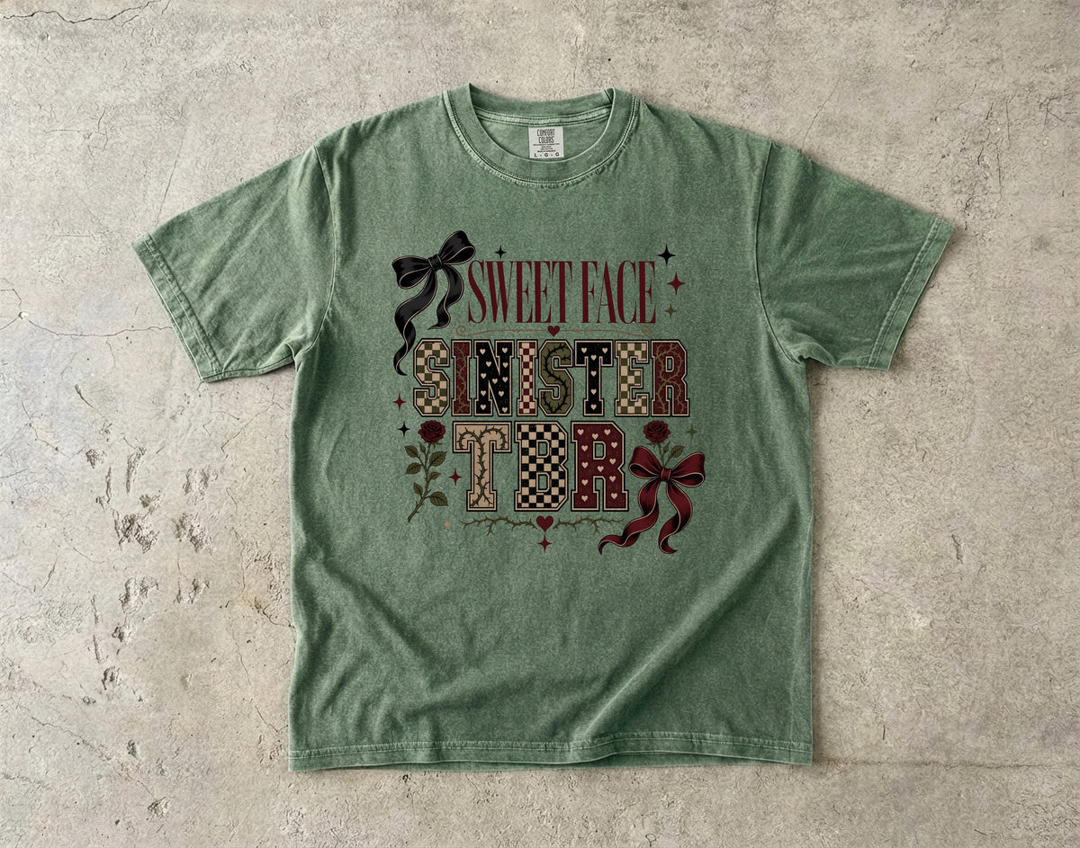 A green graphic t-shirt laid flat on a textured concrete surface. The shirt features a decorative graphic on the front with the words 'SWEET FACE SINISTER' in a patchwork pattern, accented with dark red bows and small floral details.